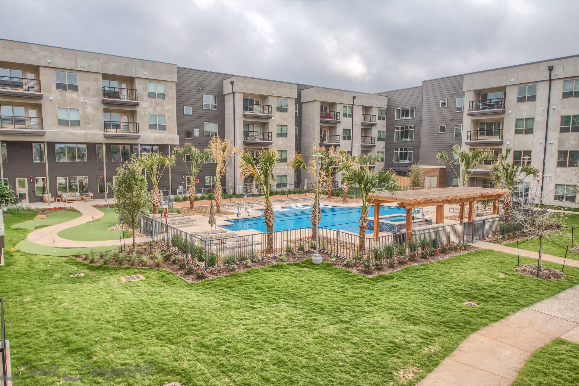 Outdoor view of Merritt Heritage Senior Village showing a large swimming pool surrounded by palm trees and a wooden pergola. The pool area is fenced and has lounge chairs. The building is a multi-story residential complex with balconies overlooking the pool and green lawn areas with walking paths.
