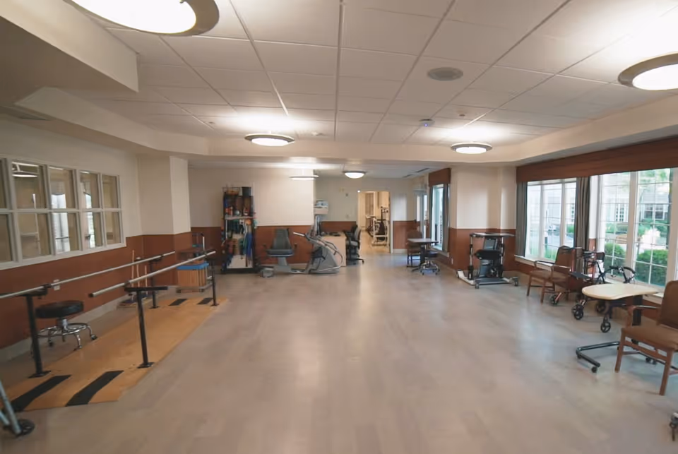 Large bright activity/therapy room with parallel bars, exercise equipment, chairs and tables along windows.