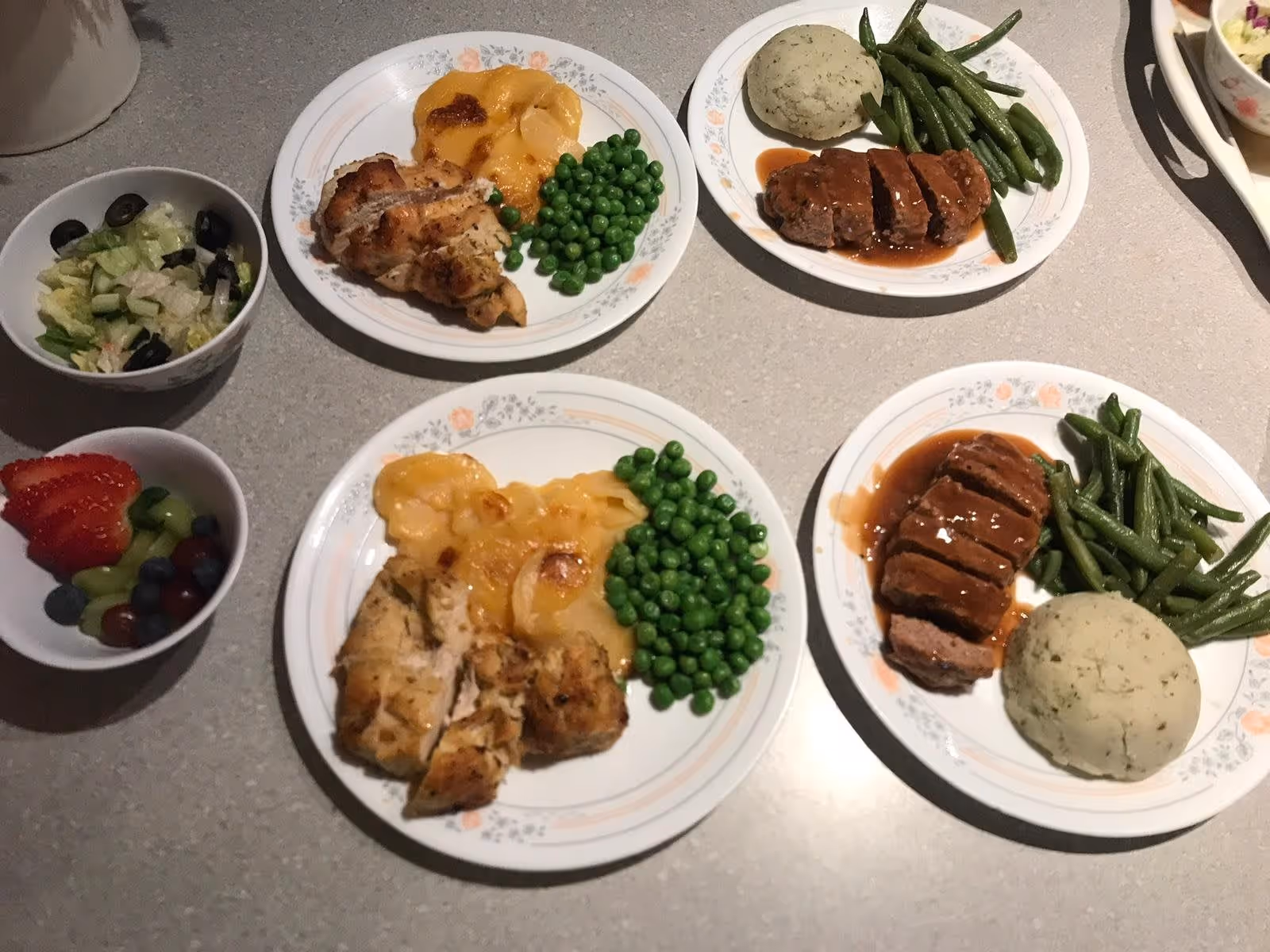 Four plates of food on a countertop, two with grilled chicken, scalloped potatoes, and green peas, and two with meatloaf, mashed potatoes, and green beans. There are also two small bowls, one with a salad containing lettuce, cucumber, and black olives, and the other with mixed fruit including strawberries, grapes, and blueberries.