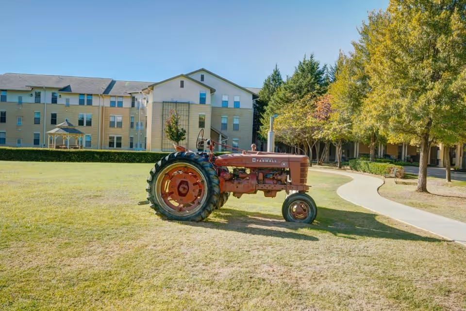 An old red Farmall tractor parked on a grassy lawn in front of a multi-story senior living facility building with a gazebo and trees nearby under a clear blue sky.