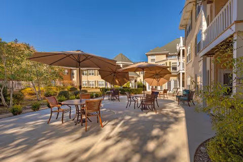 Outdoor patio area at Elison Assisted Living of Oxford with multiple tables and chairs under large beige umbrellas, surrounded by landscaped greenery and the exterior of the building under a clear blue sky.