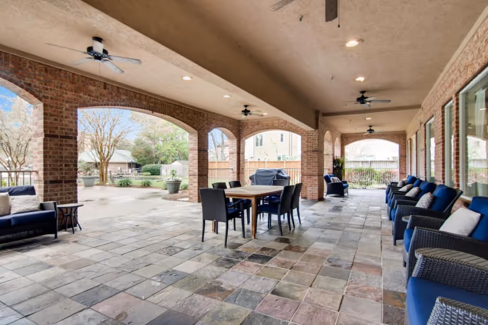 Covered outdoor patio area with stone tile flooring, brick arches, ceiling fans, and seating including blue cushioned chairs and a dining table with chairs. The patio overlooks a garden with trees and shrubs.