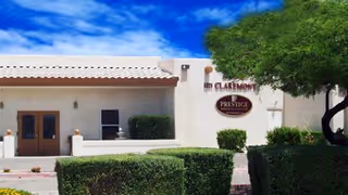 Exterior view of Claremont Assisted Living facility showing a beige building with a tiled roof, a sign reading 'Claremont Assisted Living Prestige Senior Living', green bushes, and a tree under a partly cloudy blue sky.