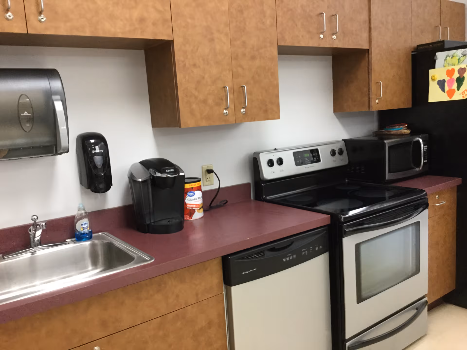 A kitchen area with wooden cabinets, a stainless steel sink with a bottle of Dawn dish soap, a Keurig coffee maker, a canister of Classic Roast coffee, a dishwasher, an electric stove with oven, a microwave, and a black refrigerator with children's artwork attached.