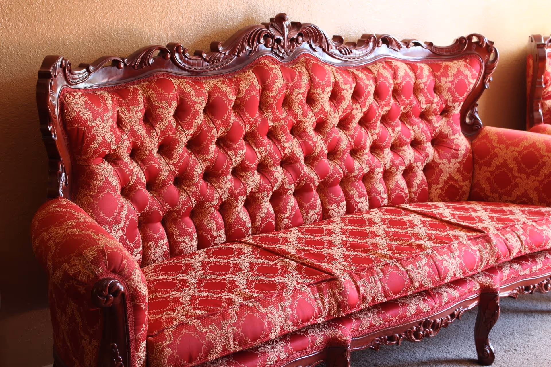 A vintage-style red upholstered sofa with intricate wooden carvings on the frame and armrests, placed against a beige wall.