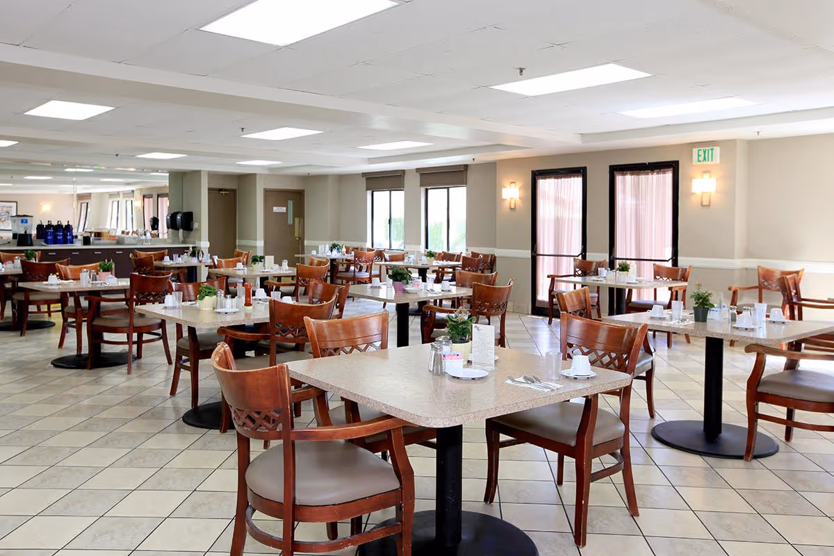 A bright and spacious dining room with multiple square tables and wooden chairs arranged neatly. Each table is set with cups, plates, utensils, and small potted plants. The room has tiled floors, large windows with blinds, and double glass doors with curtains. The walls are light-colored with wall sconces providing additional lighting.