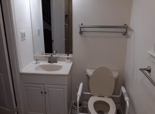 A small bathroom with a white sink vanity on the left, a mirror above the sink, and a toilet with safety rails on both sides. There is a towel rack mounted on the wall above the toilet and another towel rack on the right wall.