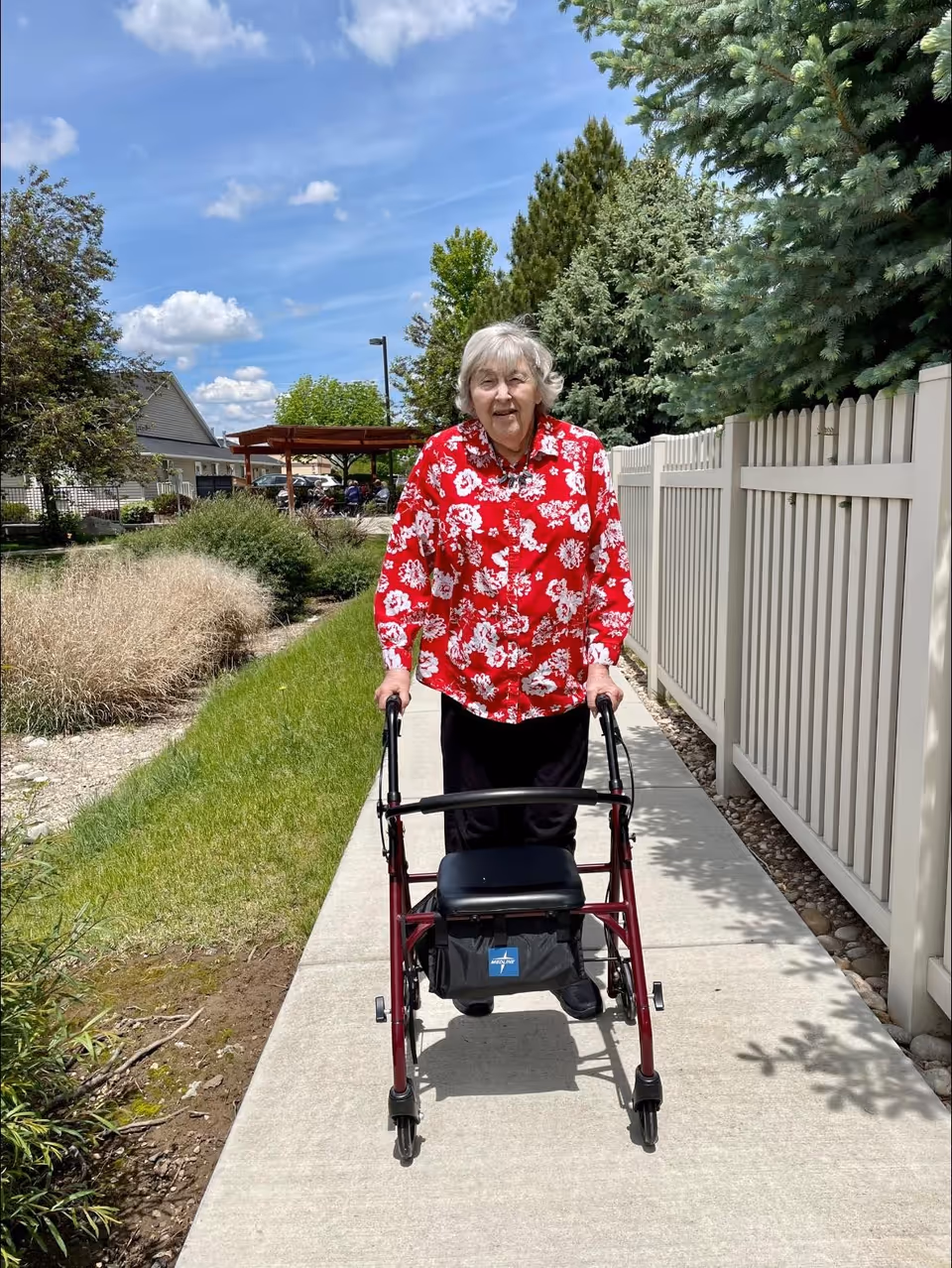 An elderly woman wearing a red floral blouse and black pants is walking outdoors on a concrete pathway using a walker. She is next to a white fence with green trees and bushes on the right side and a grassy area with shrubs on the left. The sky is blue with some clouds.