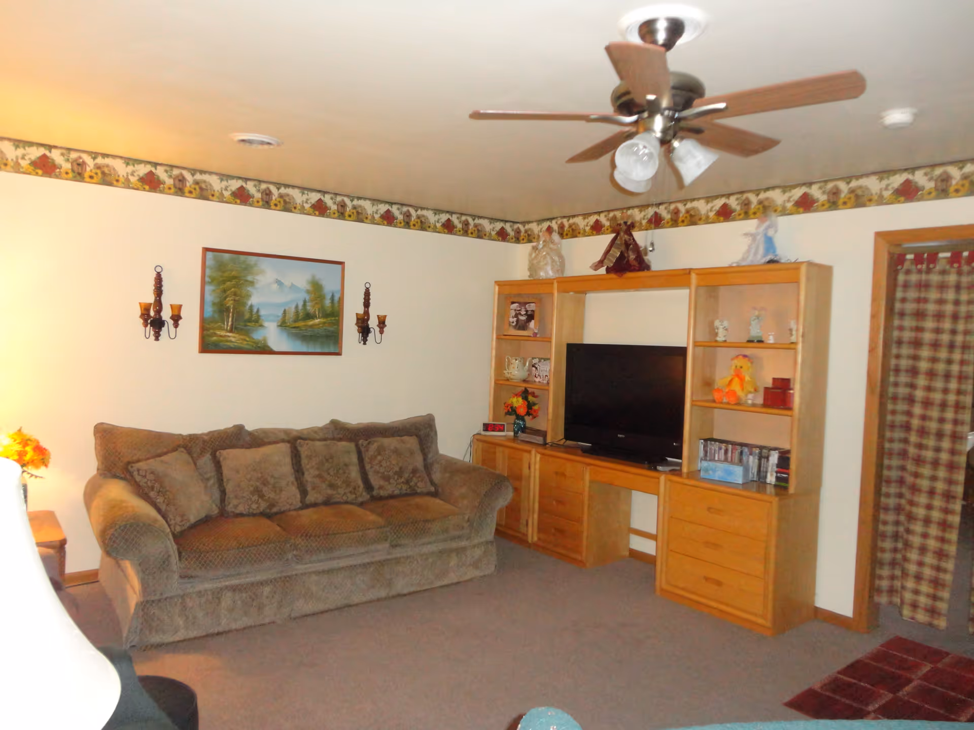 A cozy living room with a brown couch adorned with multiple cushions, a wooden entertainment center holding a flat-screen TV and decorative items, a ceiling fan with lights, and a landscape painting on the wall. The room has beige walls with a floral border near the ceiling and a plaid curtain covering a doorway.