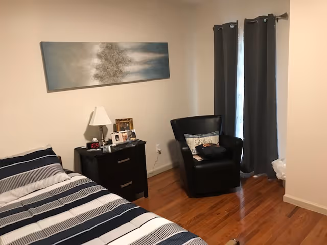 A bedroom with a bed covered in striped bedding, a black nightstand with a lamp, digital clock, and framed photos. A black armchair with a pillow and a bag is placed near a window with dark curtains. The floor is wooden and the walls are painted light beige. A rectangular abstract painting hangs above the nightstand.