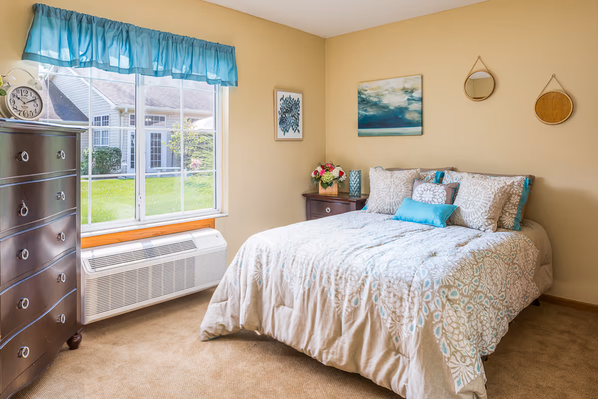 Well-lit bedroom with a double bed covered in patterned bedding, a dresser by a large window, and wall art.
