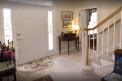 Interior view of a home's entryway featuring a white front door with sidelights, a small table with a lamp and framed pictures on the wall, a carpeted floor with a decorative rug, and a staircase with white railings leading upstairs.