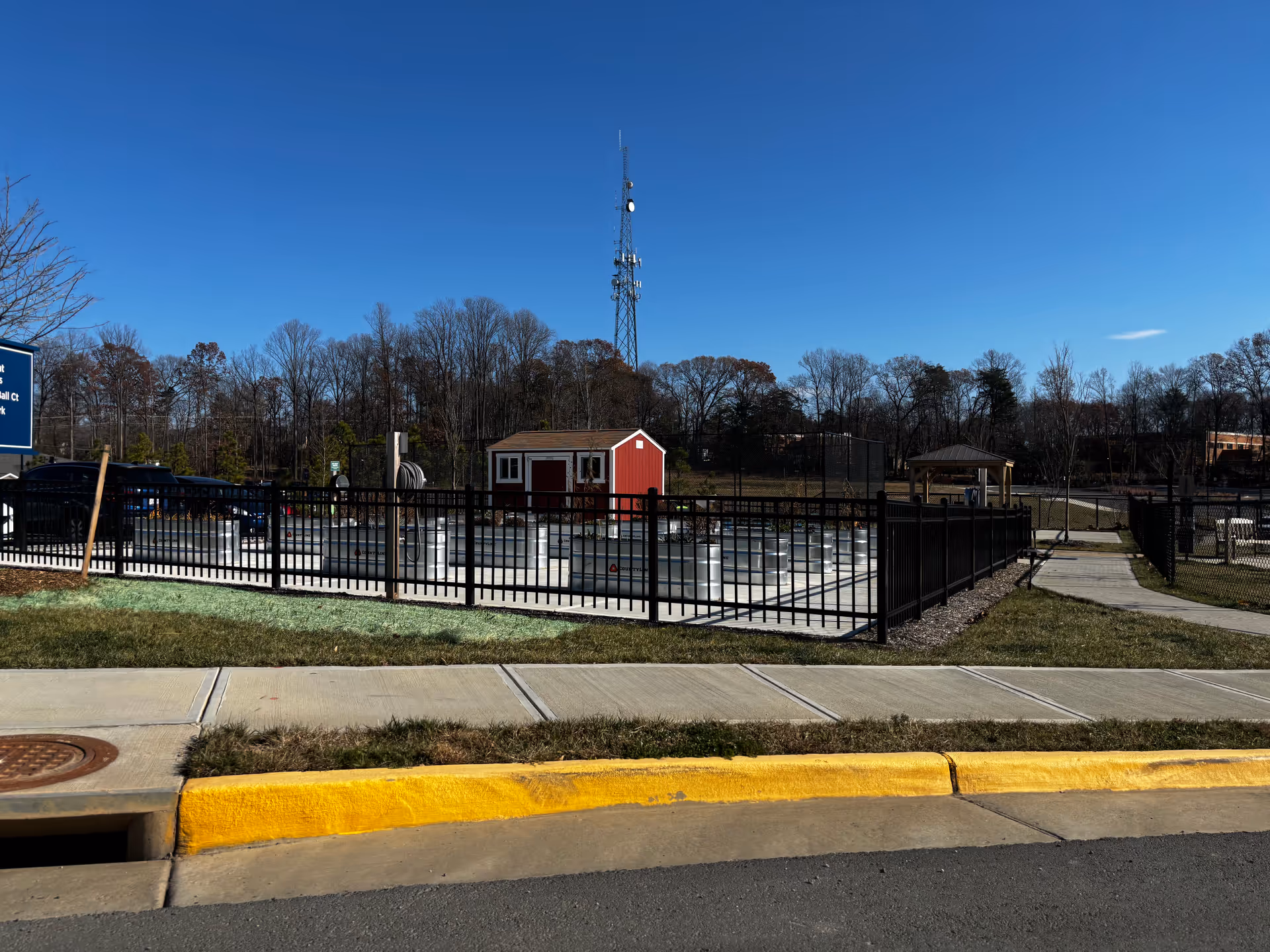 Fenced outdoor area with a red shed, gazebo, and a tall communications tower behind under a clear blue sky.