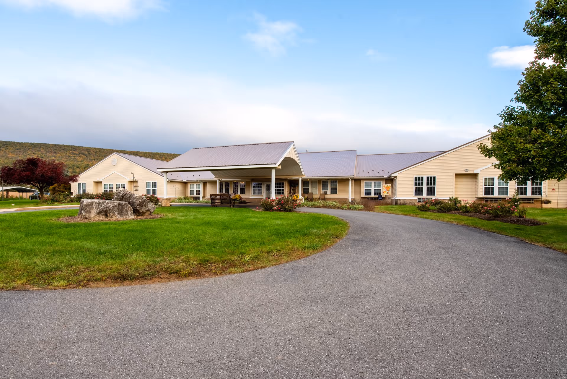 Exterior view of a single-story senior living facility building with beige siding and a metal roof. The building has a covered entrance with benches and is surrounded by green grass, trees, and a paved driveway curving in front. Hills and a partly cloudy sky are visible in the background.