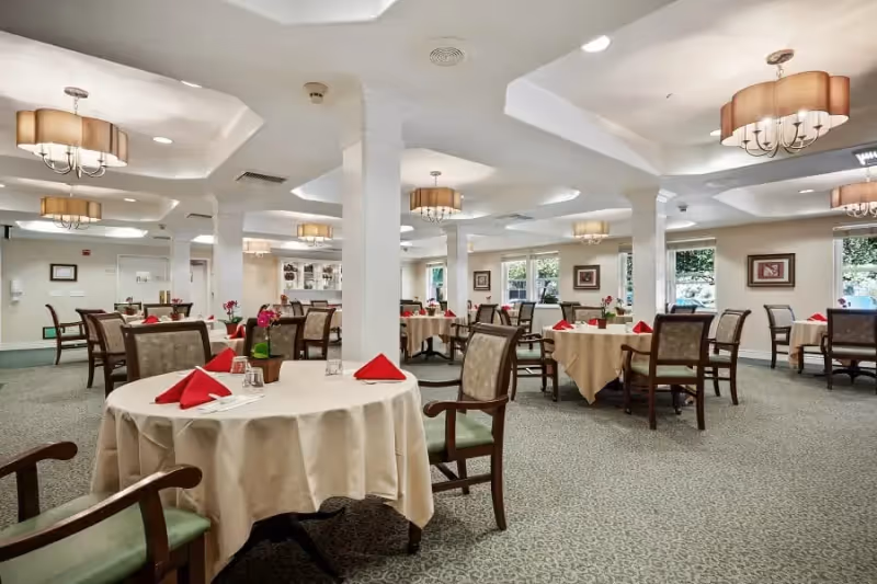 A spacious dining room with multiple round tables covered in beige tablecloths, each set with red folded napkins, glasses, and small flower arrangements. The room features carpeted floors, white columns, framed artwork on the walls, and large windows letting in natural light. Elegant ceiling light fixtures hang from the white ceiling.