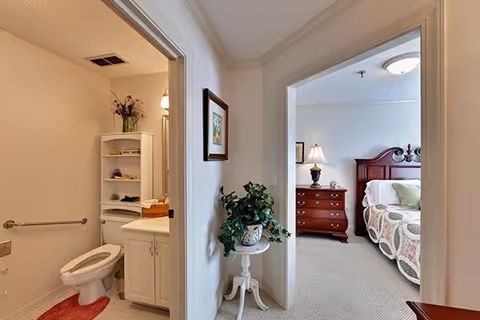 View of a hallway in an assisted living facility showing a bathroom on the left with a toilet, white cabinet, and shelves with decorative items, and a bedroom on the right with a wooden bed, nightstand with lamp, and dresser. A small table with a potted plant is placed in the hallway.