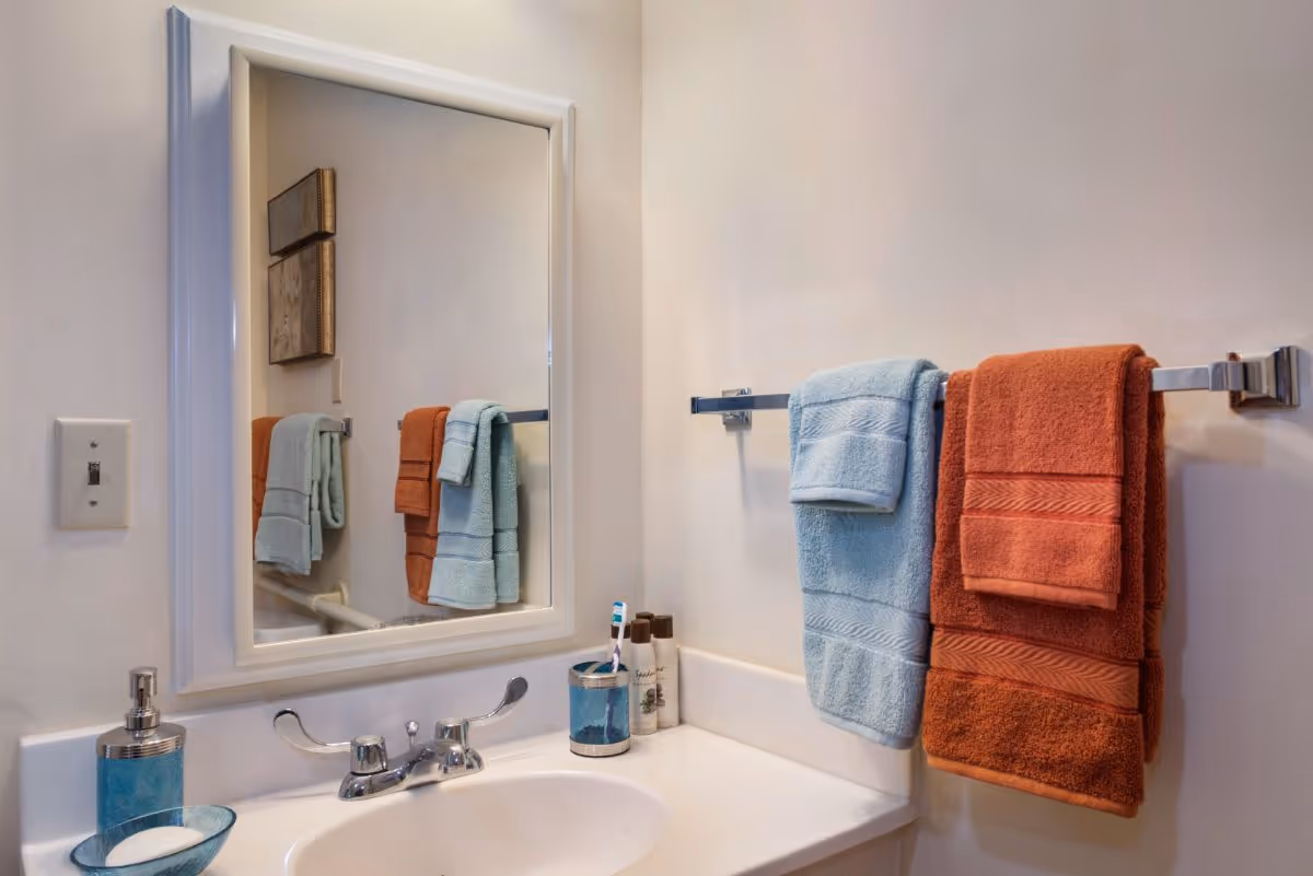 A bathroom sink area with a white countertop and a silver faucet. Above the sink is a rectangular mirror with a white frame. On the right wall, there is a towel rack holding two folded towels, one light blue and one burnt orange. On the countertop, there is a blue soap dispenser, a blue toothbrush holder with a toothbrush, and two small bottles of toiletries. The walls are painted a light beige color.