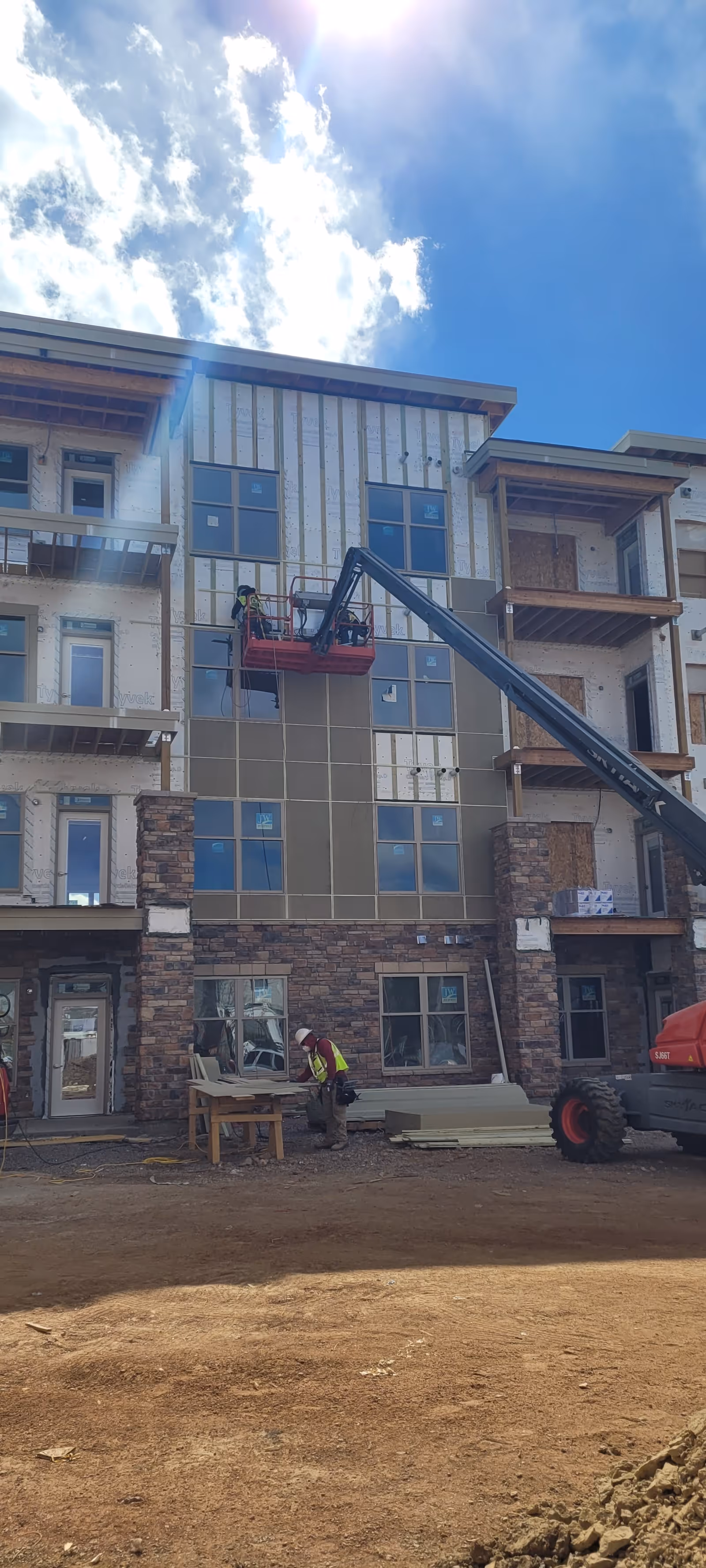 Construction workers working on the exterior of a multi-story building under construction at Aberdeen Ridge. One worker is elevated on a lift platform working on the upper facade, while another worker is on the ground near construction materials. The building features stonework on the lower level and partially installed siding on the upper levels. The sky is partly cloudy with sunlight shining through.