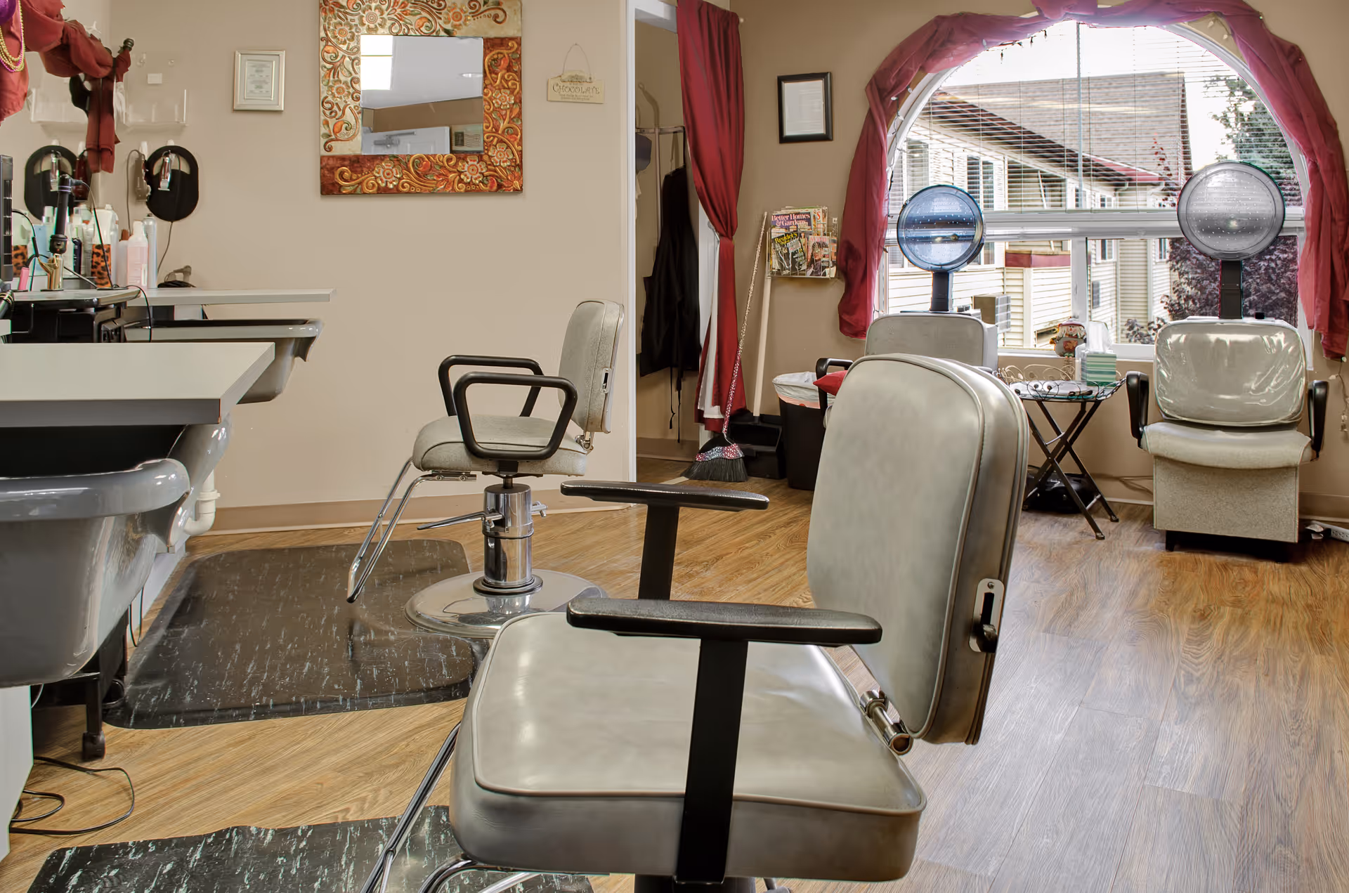 Interior of a hair salon area in a senior living facility with salon chairs, hair dryers, a mirror with a decorative frame, and a window with red curtains overlooking an exterior building.