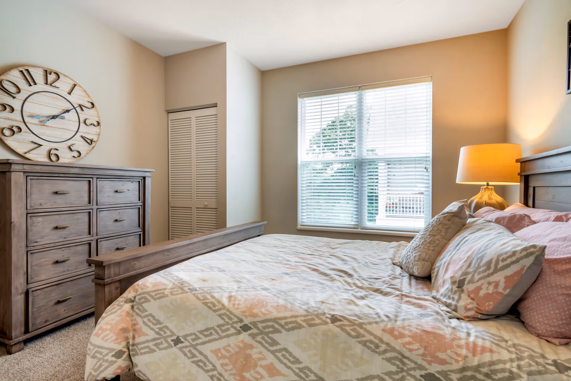 Well-lit bedroom with a wooden bed and dresser, patterned bedding, a large wall clock, and a window with blinds.