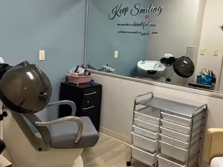 Salon-style room with a styling chair and hooded hair dryer, mirror, wash sink, storage drawers, and a 'Keep Smiling' wall decal.