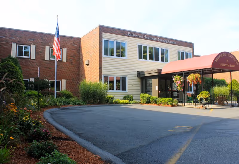 Exterior view of Briarwood Healthcare Nursing Center, a two-story building with a brick and light-colored siding facade. There is a red canopy over the entrance with hanging flower baskets and an American flag on a flagpole near the building. The surrounding area has landscaped greenery and a paved driveway.