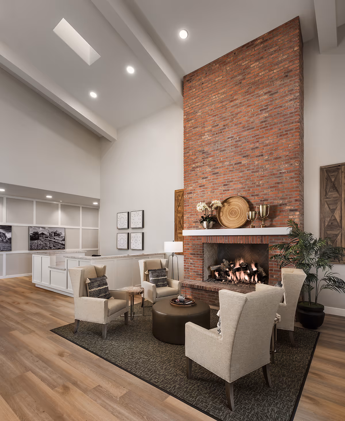 A cozy living room area with four beige armchairs arranged around a round ottoman on a dark rug. A large brick fireplace with a fire burning is the focal point, decorated with a woven basket, two gold trophies, and a potted plant on the mantel. The room has high ceilings with recessed lighting, light wood flooring, and a white reception desk in the background with black and white framed photos on the wall.