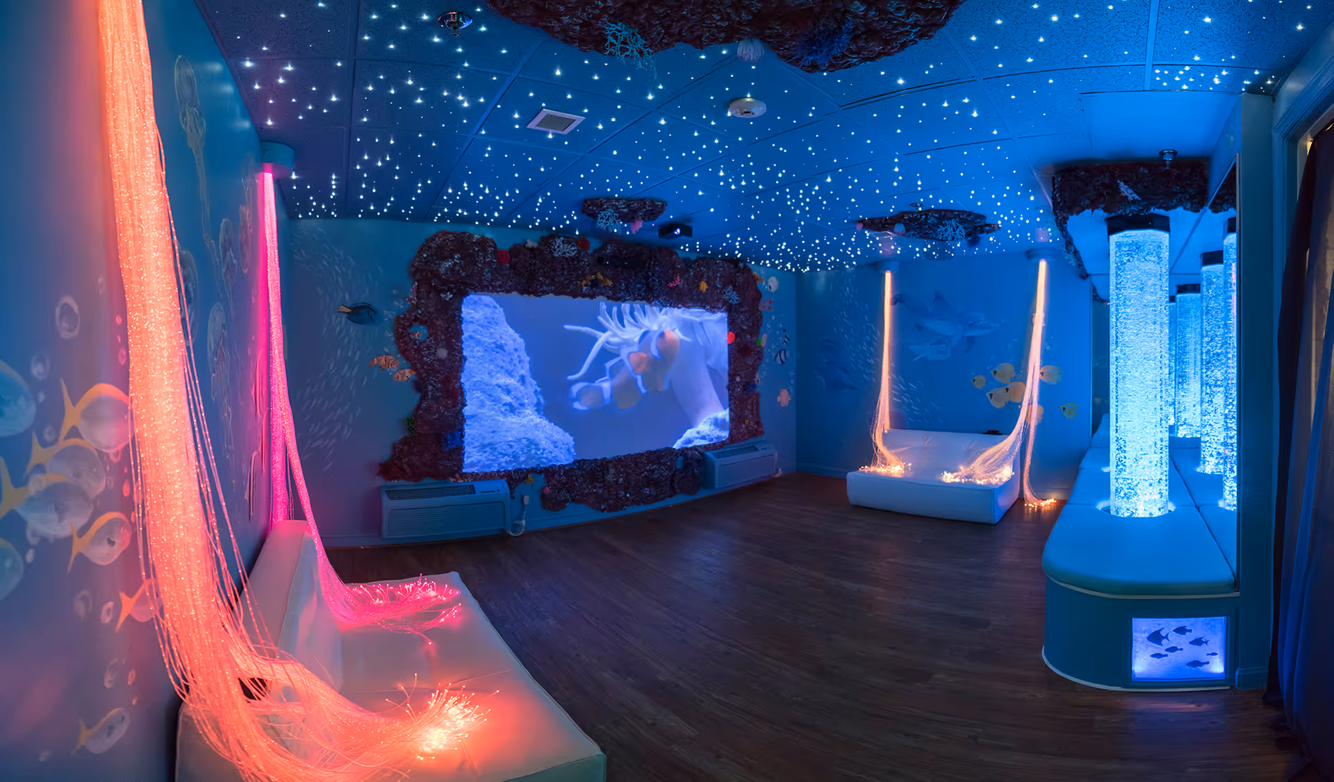 A sensory room with an underwater theme featuring glowing fiber optic lights in pink and blue, a large screen displaying underwater scenes, star-like ceiling lights, and bubble tubes along the right side. The walls are decorated with fish and coral designs, and there are white cushioned benches for seating.