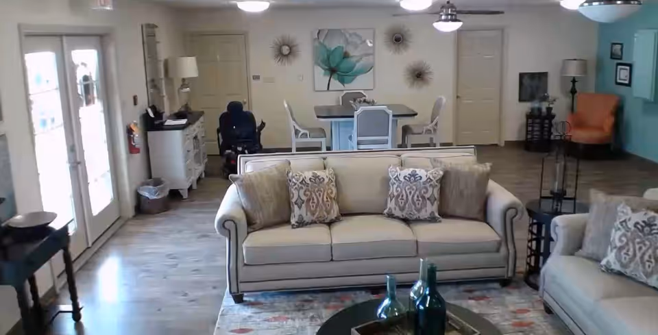 A bright and spacious living room area in an assisted living community featuring two beige sofas with decorative pillows, a round coffee table with decorative bottles, a dining table with chairs in the background, and various wall decorations including a large floral painting and sunburst wall art. There is a wheelchair near a cabinet with a lamp, and large glass doors letting in natural light on the left side.