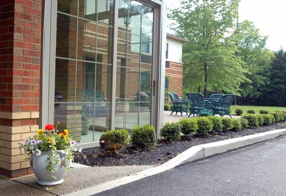Outdoor patio area at The Pines of Mount Lebanon featuring a brick building with large glass windows, a row of small green bushes along the edge of the sidewalk, a flower pot with colorful flowers, and green patio chairs and tables set on a concrete surface surrounded by trees and greenery.