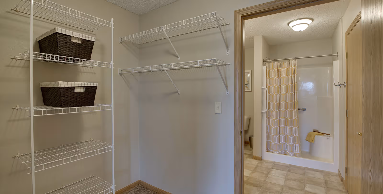 A closet area with white wire shelving and two dark woven baskets on the left side. To the right, there is an open doorway leading to a bathroom with a shower that has a yellow and white patterned curtain, a grab bar, and a towel hanging on a built-in seat. The bathroom floor is tiled and there is a ceiling light fixture.