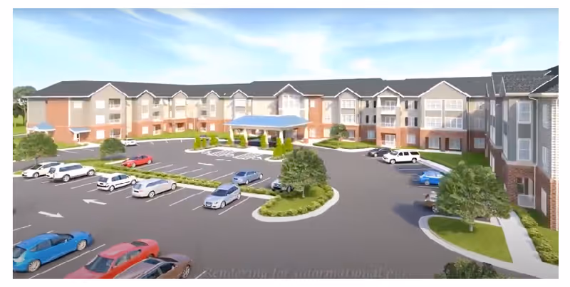 Exterior view of a large senior living facility building with multiple floors, a covered entrance, and a spacious parking lot with several cars parked. The building has a combination of brick and light-colored siding with multiple windows and landscaped greenery around the parking area.