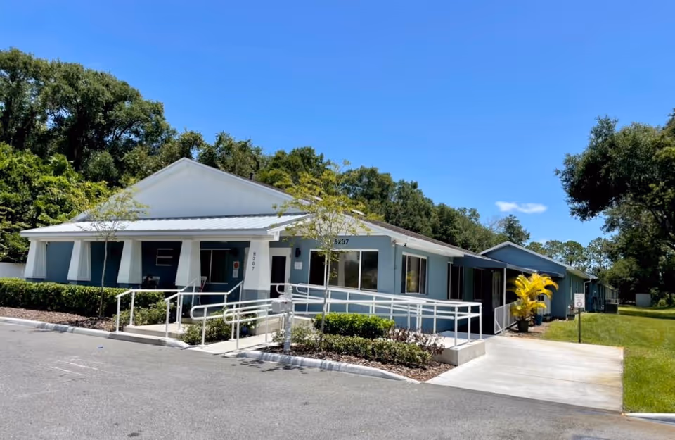 Single-story building with a white roof and blue exterior walls, featuring a wheelchair accessible ramp and surrounded by greenery under a clear blue sky.