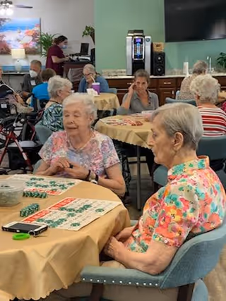 A group of elderly people sitting around tables covered with yellow tablecloths, playing bingo in a common room. Some individuals are focused on their bingo cards, while others are engaged in conversation. A staff member wearing a mask is seen in the background near a counter with a coffee machine and a television mounted on the wall.