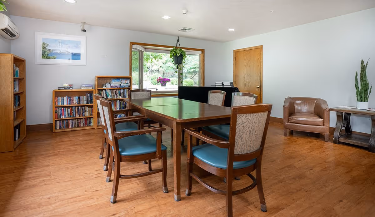 A bright room with a wooden table surrounded by six chairs with blue cushions. There are bookshelves filled with books against the wall, a large window with a view of greenery outside, a hanging plant, a brown leather armchair, and a side table with a potted plant. The floor is wooden and the walls are white.