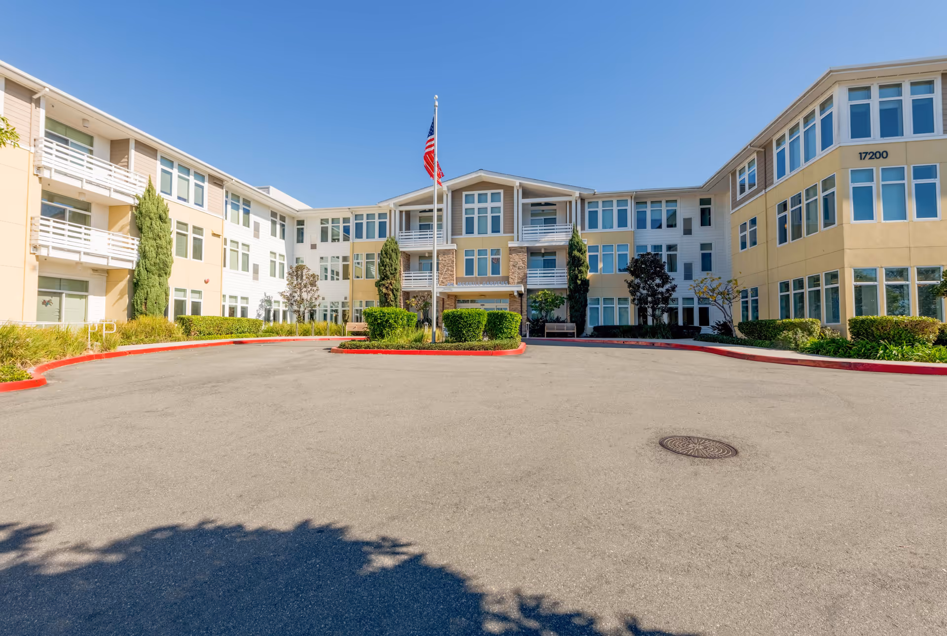 Exterior view of a three-story senior living facility building with multiple windows, balconies, and an American flag on a flagpole in front. The building is surrounded by neatly trimmed bushes and trees, with a paved driveway and red curbs in the foreground under a clear blue sky.