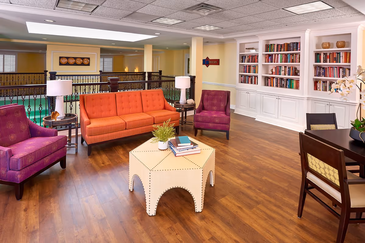 A cozy sitting area in an assisted living facility featuring an orange sofa, two purple armchairs, a white hexagonal coffee table with books and a plant, a dark wood dining table with chairs, and a built-in white bookshelf filled with books. The room has wood flooring, soft yellow walls, and a ceiling with recessed lighting. A sign on the wall indicates the direction to the library.