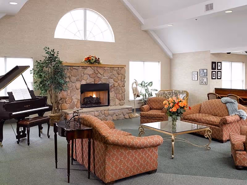 A cozy living room with a stone fireplace, a grand piano, several upholstered sofas and armchairs arranged around a glass coffee table with a vase of flowers, large windows letting in natural light, and framed pictures on the walls.