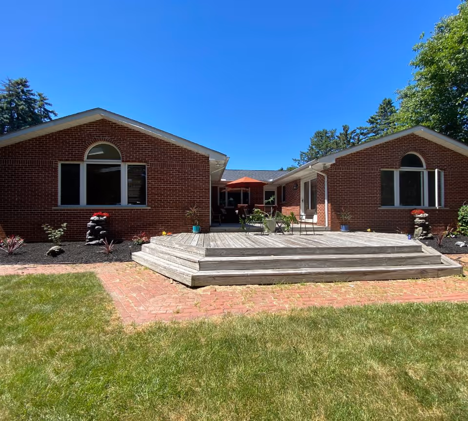 Rear exterior of a single-story red brick building with a wooden deck, patio furniture and umbrella overlooking a grassy lawn.