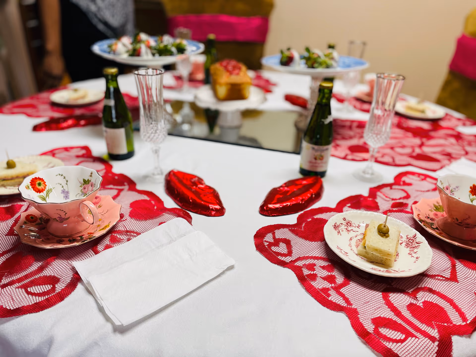 A decorated dining table with floral teacups, small sandwiches on plates, mini champagne bottles, crystal glasses, and red lace heart placemats.