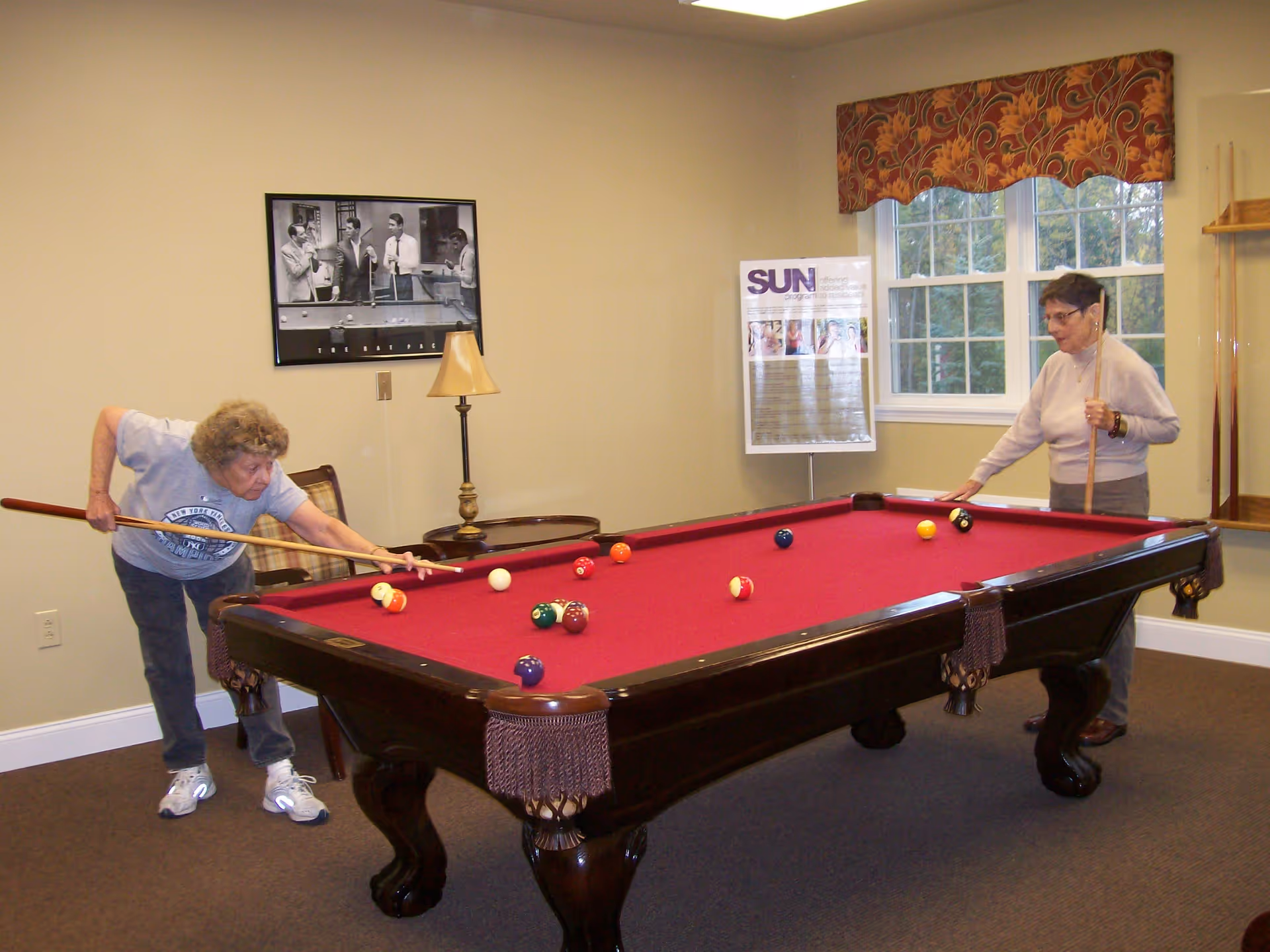Two elderly women playing pool on a red felt pool table in a well-lit room with beige walls. One woman is taking a shot while the other watches, holding a cue stick. There is a lamp on a small table against the wall, a framed black and white photo, and a window with a patterned valance.