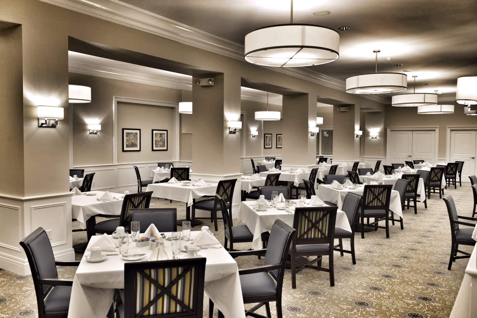 A spacious, elegant dining room with multiple tables covered in white tablecloths, each set with napkins, glasses, and cups. The room features beige walls, decorative wall sconces, large round ceiling lights, and framed artwork on the walls. Dark wooden chairs with cushioned seats surround the tables, and the carpet has a subtle patterned design.
