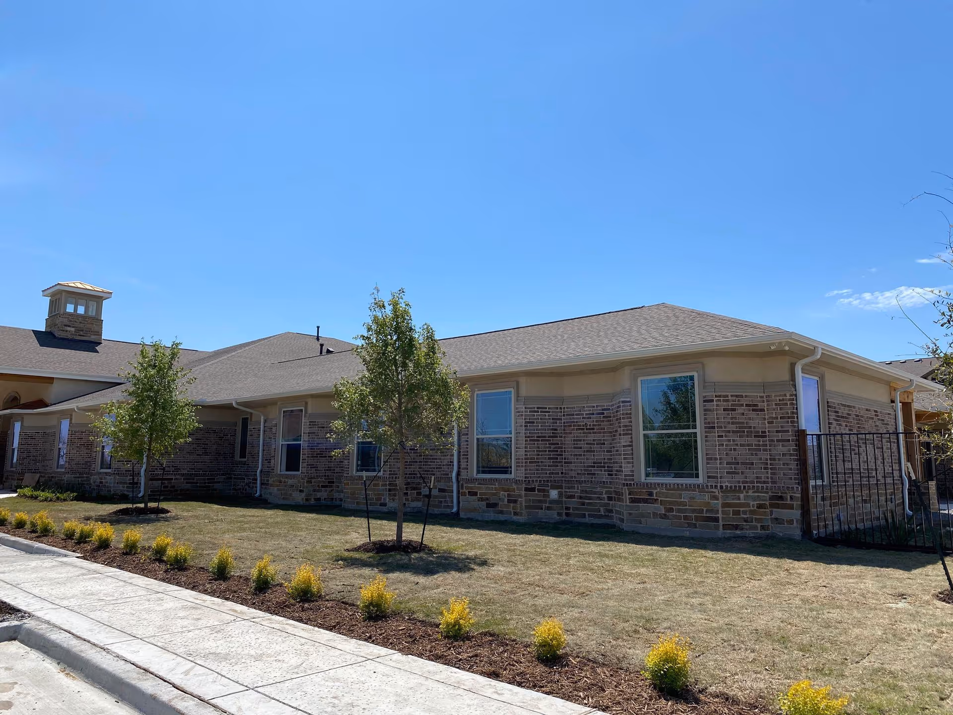 Exterior view of a single-story brick building with multiple windows, a sloped roof, and a small landscaped area with young trees and shrubs under a clear blue sky.