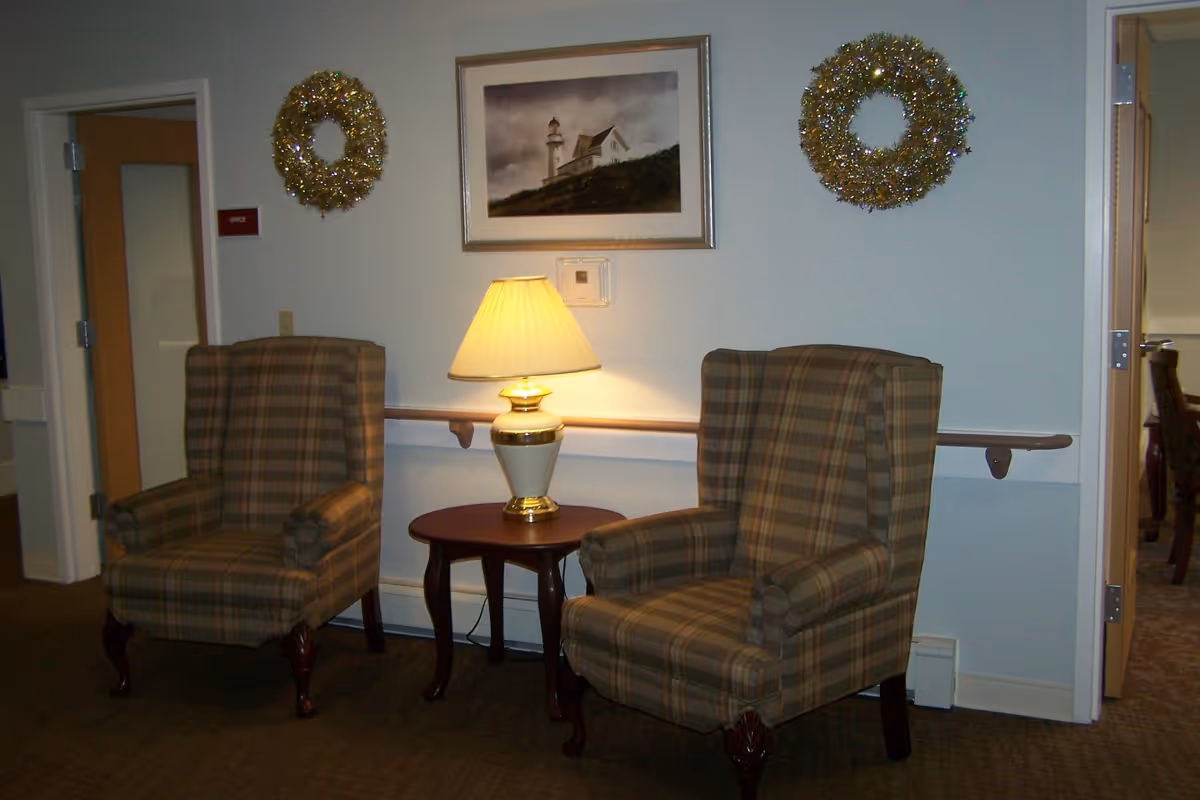 Two plaid upholstered armchairs with wooden legs positioned on either side of a small wooden table with a lit table lamp. Above the table hangs a framed picture of a lighthouse. Two decorative wreaths are mounted on the wall, one on each side of the picture. The setting appears to be an indoor common area or lounge with doors visible on either side.