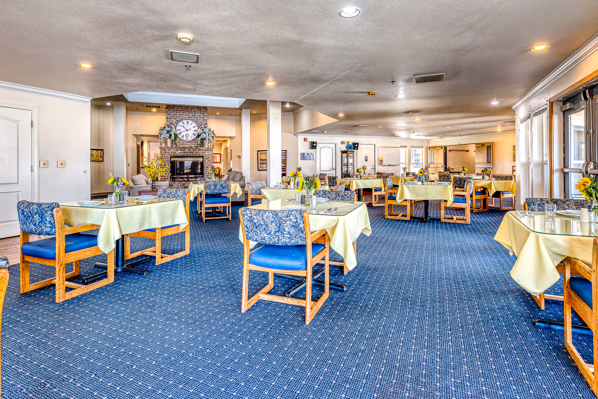 Spacious, brightly lit dining room with multiple tables set with yellow tablecloths, wooden chairs, and a fireplace in the background.