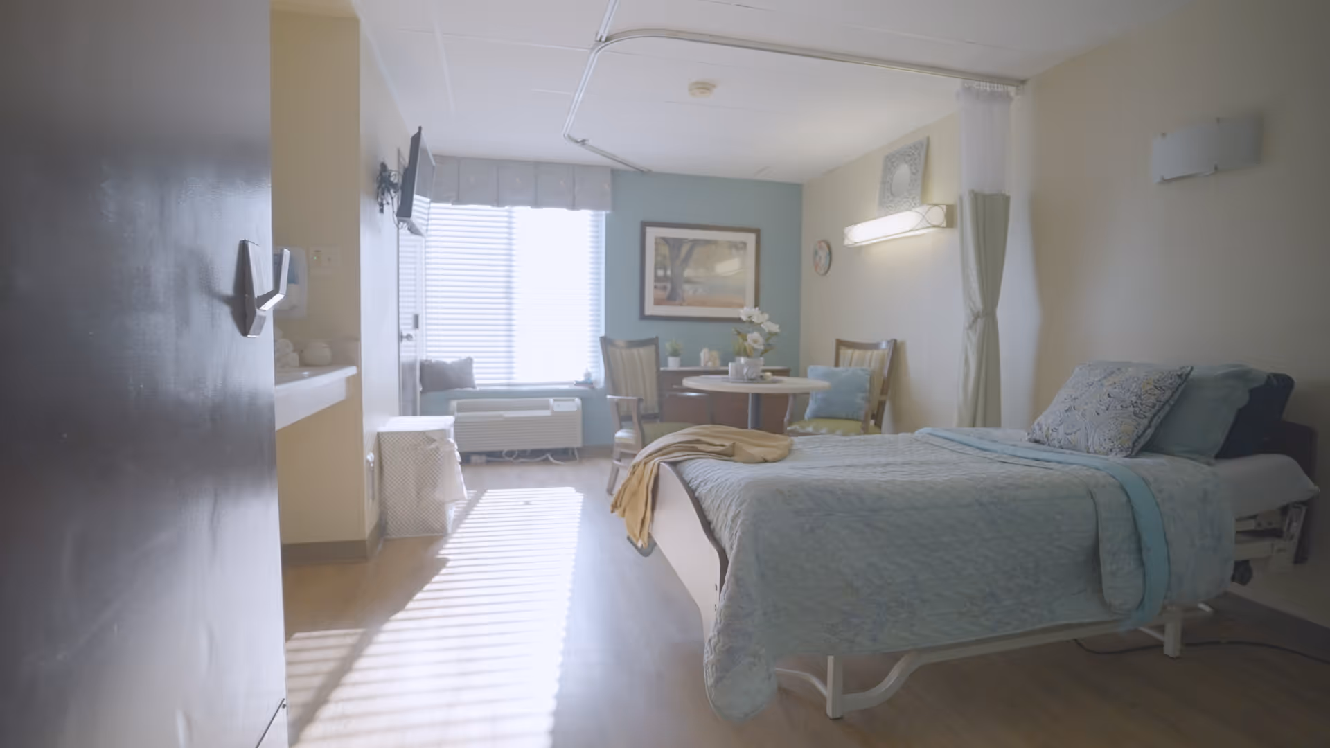 A bright and clean bedroom in a senior care facility with a single bed covered in light blue bedding, a small round table with two chairs, a window with blinds letting in sunlight, a wall-mounted TV, and a small sink area near the door.