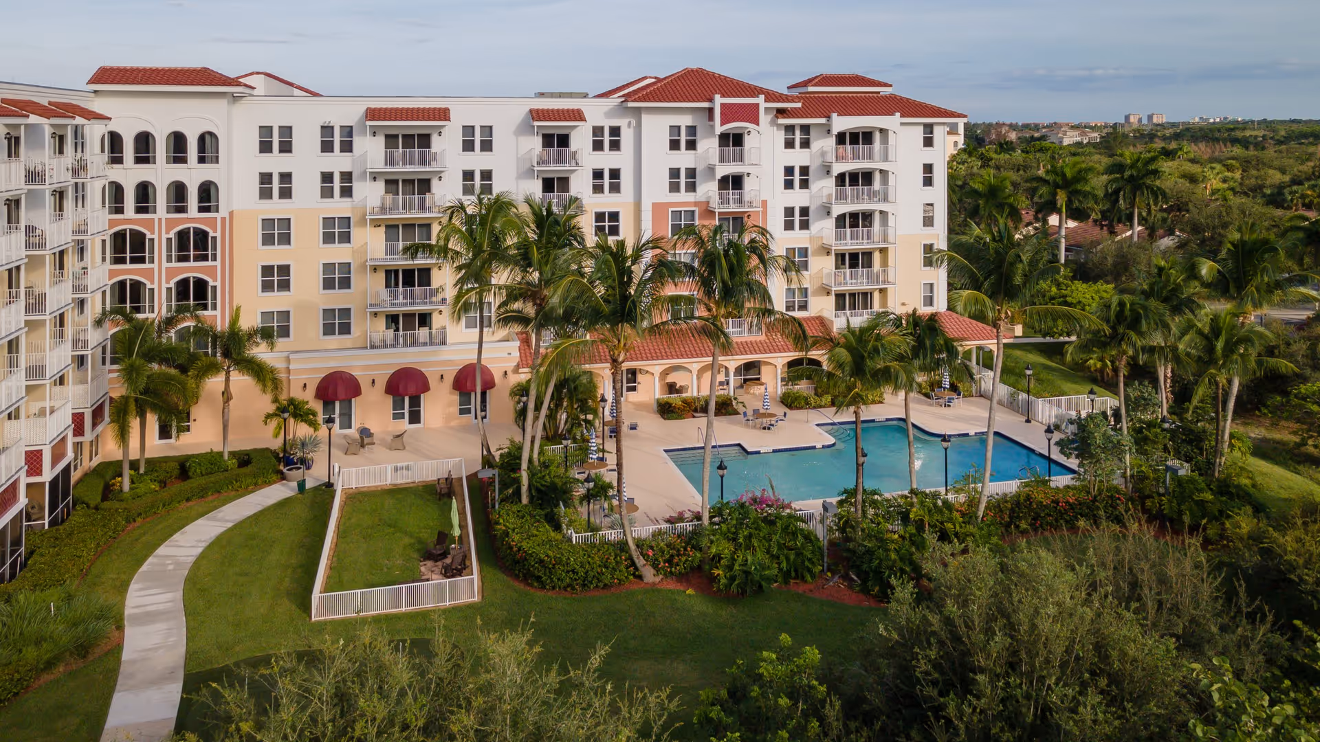 Exterior view of a multi-story senior living facility named Mangrove Bay with a swimming pool surrounded by palm trees and landscaped greenery. The building has balconies and red-tiled roofs under a clear sky.