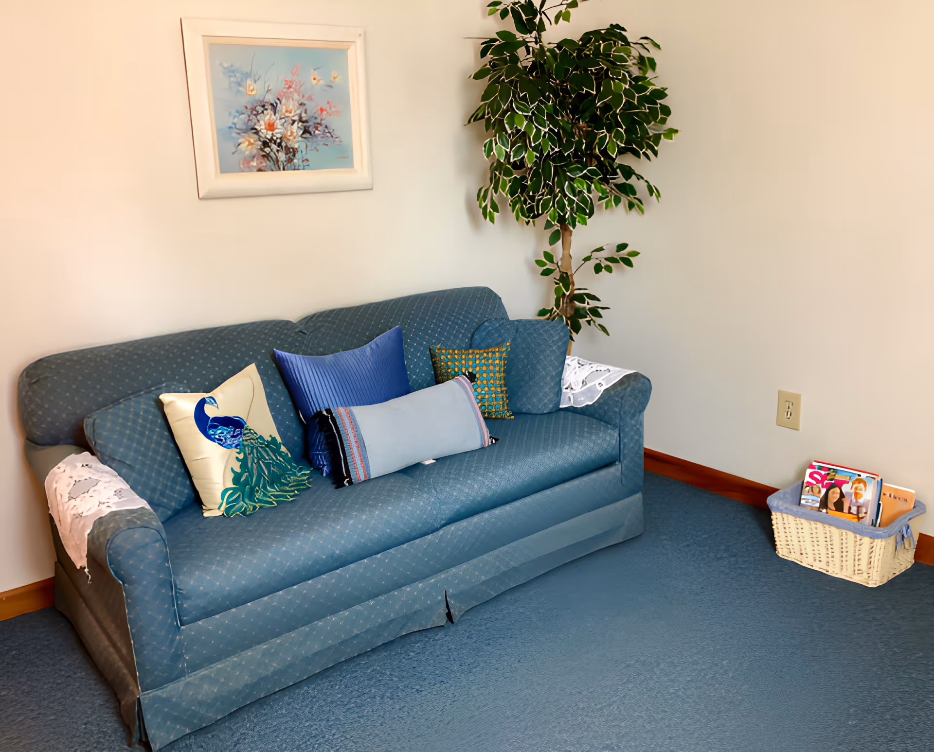 Blue upholstered sofa with decorative pillows, a tall potted plant, framed artwork above, and a basket of magazines on the floor in a small seating area.