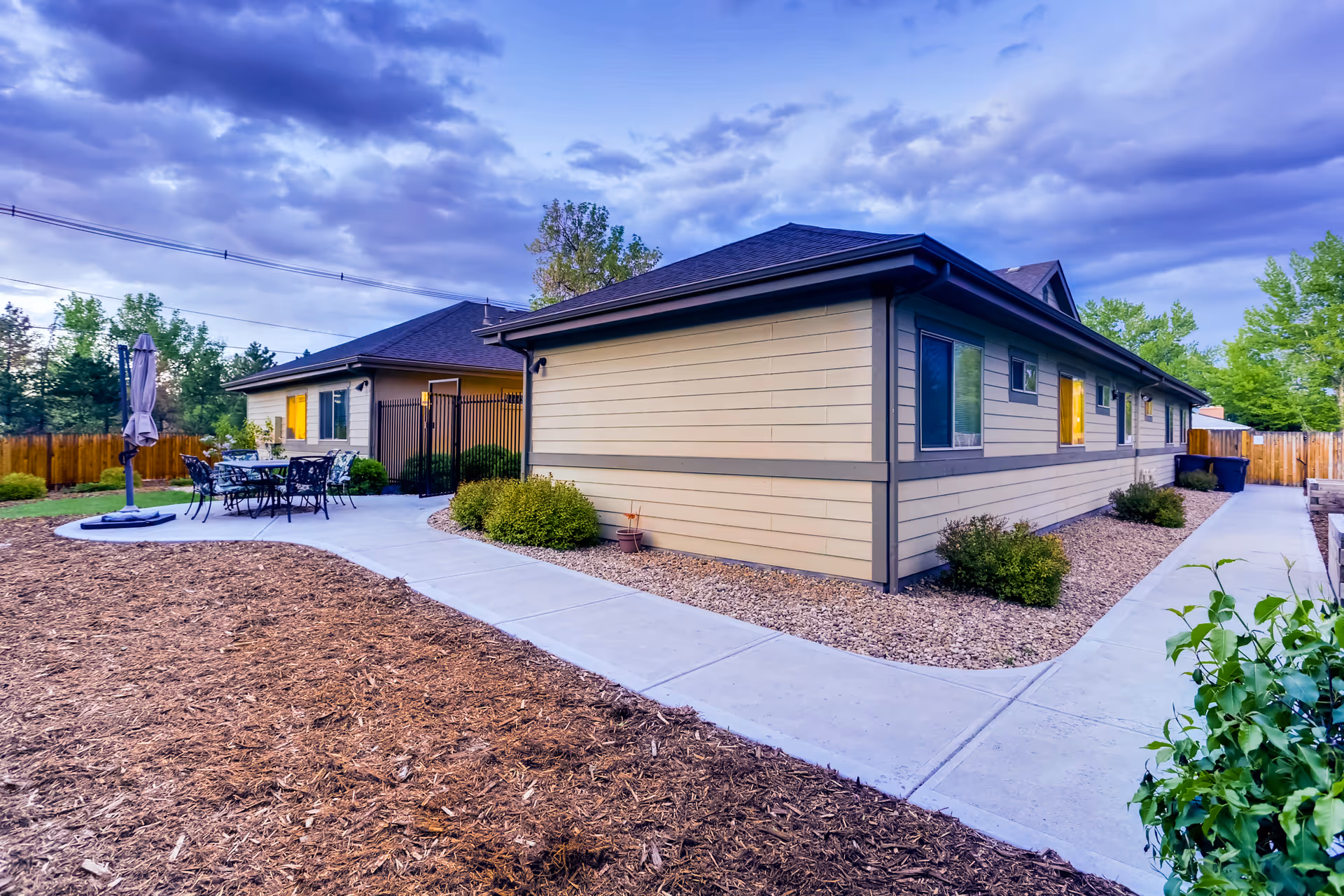 Exterior view of a single-story assisted living building with a paved walkway, patio seating and landscaped grounds under a cloudy sky.