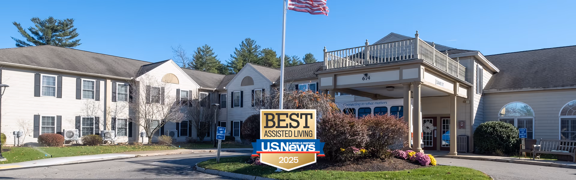 Exterior view of Benchmark Senior Living at Nashua Crossings, showing a two-story building with white siding and black shutters. There is a covered entrance with columns and a sign in front that reads 'BEST ASSISTED LIVING U.S. News 2025'. The area is landscaped with bushes and flowers, and there are trees in the background under a clear blue sky.